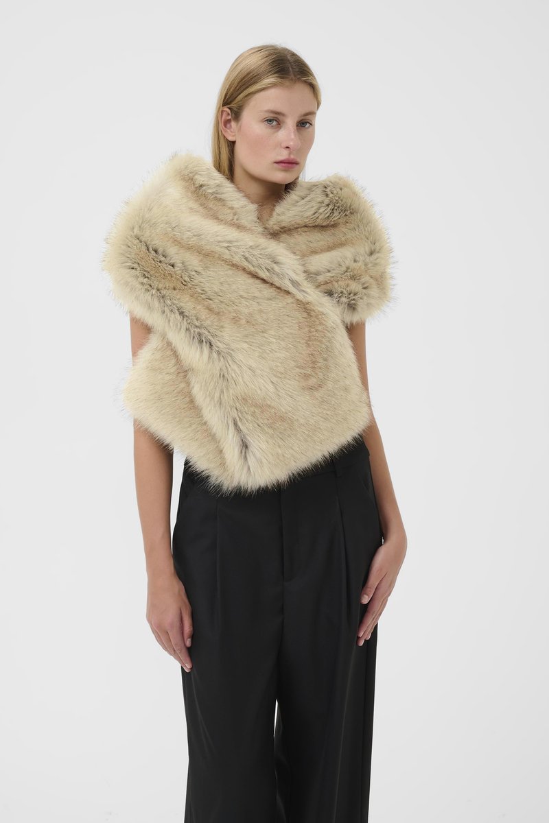 Donna che indossa uno scialle di pelliccia beige drappeggiato su spalle nude e pantaloni a gamba larga neri a vita alta, in piedi contro uno sfondo semplice.