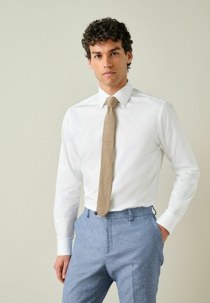 Mann mit lockigem Haar, der ein weißes Hemd, eine beige gestrickte Krawatte und hellblaue maßgeschneiderte Hose vor neutralem Hintergrund trägt.