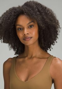 Pruun, ilma varrukat kleit v-kujulise kaelusega, sileda tekstuuri ja lihvitud kujuga. Kurruline, tume juuksed raamivad nägu neutraalses taustas.