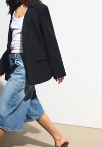 Veste blazer noire surdimensionnée avec revers crantés et poches avant, associée à un pantalon culotte en denim bleu clair et des sandales marron, sur un fond uni.