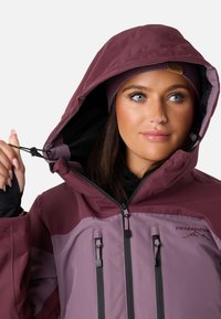 Giacca da donna color bordeaux e viola con cappuccio regolabile, cerniere e dettagli neri. Tessuto testurizzato con logo del marchio in evidenza.