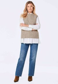 Beige gestricktes ärmelloses Oberteil, das über einem weißen Hemd mit Knopfleiste getragen wird, kombiniert mit blauen geraden Jeans und braunen Knöchelstiefeln.