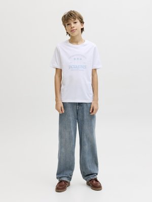 Vaaleanruskeatukka poika, joka on pukeutunut valkoiseen Jack & Jones -t-paitaan, väljät siniset farkut ja ruskeat kengät, seisoo yksinkertaista vaaleaa taustaa vasten.