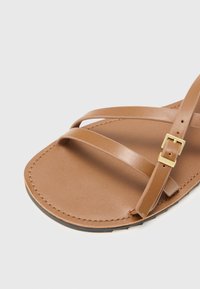 Bruine platte sandaal met dunne, kruislings straps van soepel leer. Verstelbare gespsluiting in goudkleur en een gestructureerde zool voor grip.