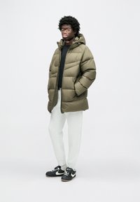 Olivno zelena puhasta bunda s kapuco, z zadrgo in stranskimi žepi; kombinirana s sivimi športnimi hlačami in črnimi športnimi copati.