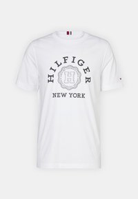 Camiseta de algodón blanca con cuello redondo, mangas cortas y un gran logo en azul marino con el texto "HILFIGER" y "NEW YORK" en un diseño circular.