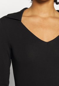 Femme portant un haut noir à manches longues avec un décolleté en V profond et un large col, laissant apparaître une partie de son cou et du haut de sa poitrine.