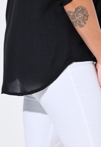 Schwarze, durchsichtige Hemdbluse mit abgerundetem Saum, kombiniert mit eng sitzenden weißen Jeans. Enthält ein dekoratives Blumentattoo am Unterarm.