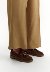 Mocassini in suede marrone con nappine, caratterizzati da dettagli in metallo, abbinati a pantaloni larghi color marrone chiaro.