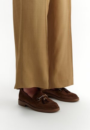 Mocassini in suede marrone con nappine, caratterizzati da dettagli in metallo, abbinati a pantaloni larghi color marrone chiaro.