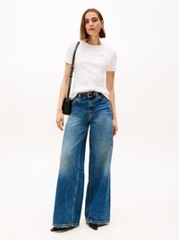 T-shirt branco de mangas curtas com um pequeno logotipo, combinado com jeans azuis de perna larga. Os jeans têm uma textura desbotada e bainha crua, finalizados com sapatos de salto pretos.