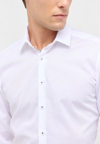 Camicia bianca con colletto classico, tessuto a trama liscia e chiusura anteriore a cinque bottoni con accenti metallici.