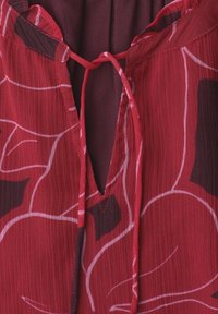 Blouse rouge avec motifs abstraits de feuilles blanches et rouge foncé, avec une encolure ajourée devant et de fines cordelettes à nouer.