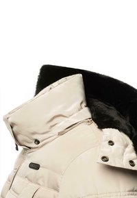 Chaqueta acolchada beige con un cuello de piel sintética negra, que cuenta con cierre de cremallera, botones a presión y diseño acolchado. Textura suave y material duradero.