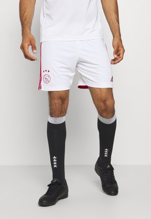 Mężczyzna noszący białe spodenki piłkarskie Ajax, czarne wysokie skarpetki w gwiazdy oraz czarne korki Adidas, stojący na jednolitym tle.