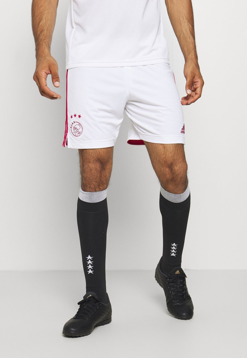 Homme portant un short de football blanc de l'Ajax, des chaussettes noires hautes avec des étoiles, et des crampons Adidas noirs, debout sur un fond uni.