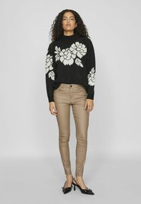 Črn pulover z belim cvetličnim vzorcem, bež oprijet hlačami in črnimi čevlji s petami. Model stoji ob enobarvni ozadju.
