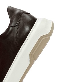 Zapatilla de cuero marrón oscuro con punta redondeada, suela de goma blanca y acentos texturizados. Presenta un diseño sin costuras y costuras sutiles.