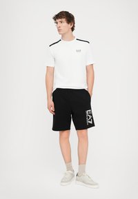 White short-sleeve t-shirt with black shoulder accents and EA7 logo, paired with black shorts featuring a white EA7 print, worn with light sneakers.