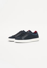Sneakers blu scuro con tessuto testurizzato, dettagli in pelle liscia, suola intermedia bianca e rivestimento rosso vicino alla caviglia, dotati di allacciatura tradizionale.