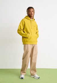 Hoodie en polaire jaune avec une poche kangourou, associé à un pantalon large beige. Modèle portant des baskets blanches et grises. Design uni.