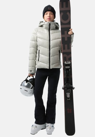 Puffige silberne Skijacke, schwarze Skihose, weiße Skischuhe, schwarze Mütze, hält einen schwarzen Ski mit roten Akzenten und weißen Helm.