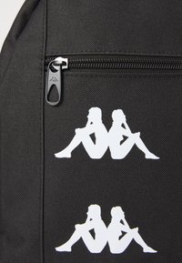 Zwarte stoffen tas met twee witte logo's die twee zittende figuren rug aan rug tonen, nabij een rits met een metalen trekker.