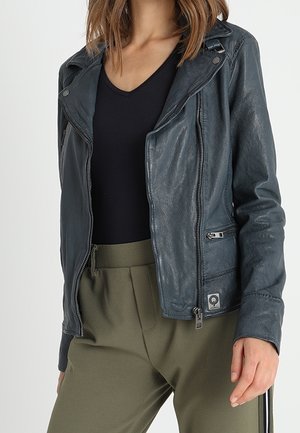 Frau trägt eine dunkelgraue Lederjacke über einem schwarzen V-Ausschnitt-Oberteil und olivgrünen Hosen mit einer Reißverschlusstasche an der Seite der Jacke.