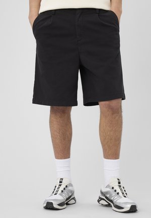Homme portant un short noir jusqu’aux genoux, des chaussettes blanches et des chaussures de sport blanches et grises, debout les mains dans les poches.
