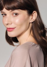 Błyszczące kolczyki wkrętki o kwadratowym kształcie, z przezroczystym kamieniem osadzonym w metalach w kolorze srebrnym. Gładki, teksturowany wierzch z miękkiego beżowego materiału.