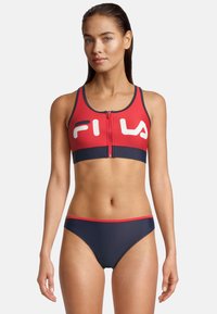 Rood en marineblauwe bikini-top met ritssluiting en wit logo, bijpassende marineblauwe bikinionderkant, gladde textuur, sportieve snit met minimale bedekking.