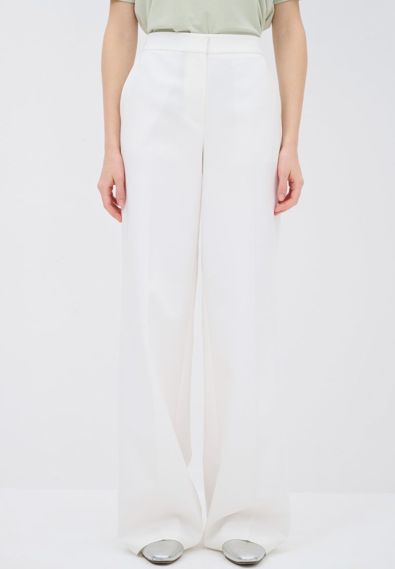 Person mit weißen, weit geschnittenen Hosen und silbernen, spitzen Schuhen, die vor einem schlichten weißen Hintergrund steht.