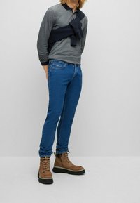Man som bär blå jeans, bruna snörstövlar, grå långärmad polo och en marinblå sweatshirt knuten runt bröstet, stående mot en neutral bakgrund.