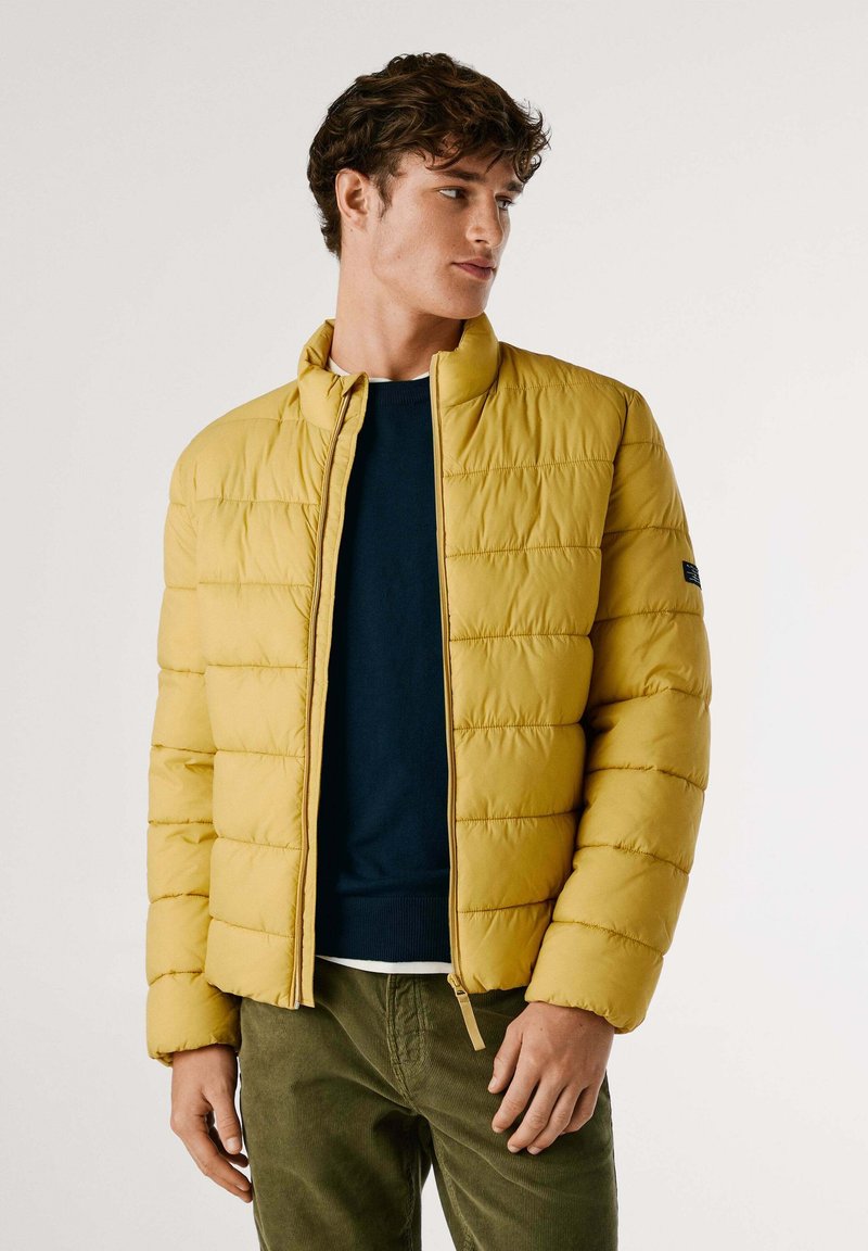 Chaqueta amarilla acolchada con cuello alto, con costuras horizontales y cierre de cremallera. Combinada con un suéter oscuro y pantalones verdes.