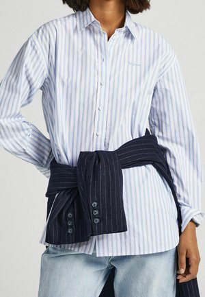 Henkilö, jolla on päällään vaaleansininen ja valkoinen pystyraitainen napillinen paita ja jonka vyötärön ympärillä on sidottu tumma ohuin raidoin kuvioitu bleiseri.