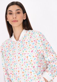 Let strikket bomberjakke med hvid bund, der har et farverigt blomstermønster i pink, orange, grøn og blå. Ribbet krave og manchetter.