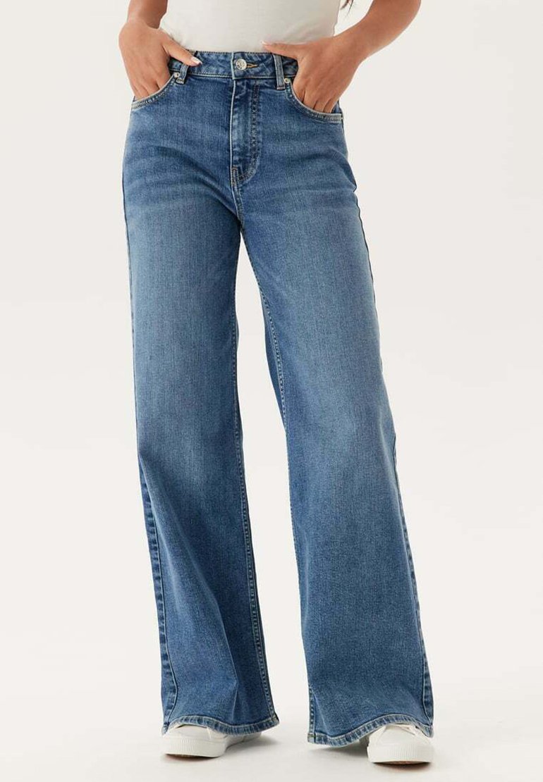 Kvinde iført højtsiddende, vidde ben blå jeans med hænderne i lommerne og hvide sneakers mod en ensfarvet baggrund.