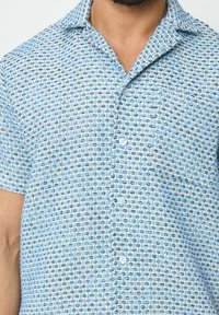 Chemise à manches courtes avec boutons, en tissu texturé de couleur bleu clair, ornée d'un motif avec de petits accents noirs et blancs.