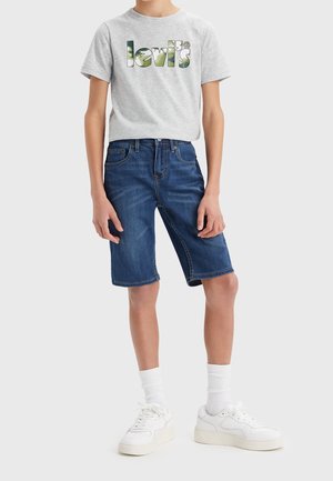 Dziecko ubrane w szarą koszulkę Levi’s z zielonym logo, niebieskie dżinsowe szorty sięgające kolan, białe skarpetki oraz białe trampki, stojące na tle białego tła.