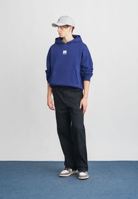 Felpa blu con tasca frontale, cappellino grigio, pantaloni neri e sneakers. La felpa presenta un piccolo patch con logo sul petto.