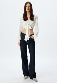 Blusa bianca cropped con dettaglio annodato davanti, abbinata a jeans a palazzo blu scuro. La blusa ha maniche lunghe con polsini elasticizzati.
