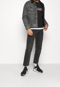 Chaqueta de mezclilla con forro negro, camiseta negra con el texto "CALVIN", jeans gris oscuro y zapatillas Nike negras con acentos blancos.