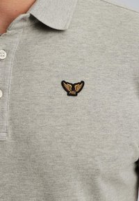 Camisa polo gris con un acabado texturizado, que presenta un emblema alado en negro y dorado en la parte izquierda del pecho, y tres botones blancos en el cuello.