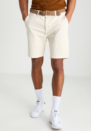 Persoon die beige korte broek draagt met een geweven bruine riem, witte sokken, witte Adidas schoenen en een ingestopt bruin shirt, handen in de zakken.