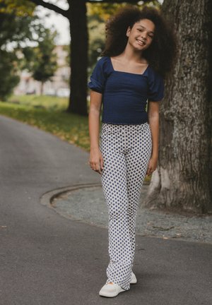 Blue smocked short-sleeve top paired with white patterned flared pants and white shoes; poses on a tree-lined walkway.