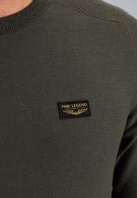 Maglione lavorato a maglia verde oliva con collo e spalle a coste. Presenta un'etichetta rettangolare nera con testo dorato e grafica di ali.