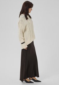 Kvinde iført en cremet kabelstrikket sweater, lang mørkebrun nederdel, sorte slingback hæle og et sort armbånd, der står i profil.