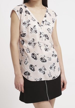 Woman wearing a sleeveless light pink blouse with black floral print and a V-neck, paired with a black skirt with a front zipper detail.