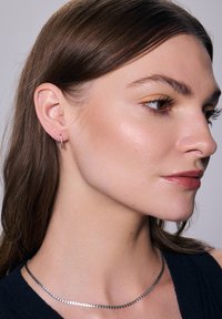 Srebrna kolczyk w kształcie obręczy na modelce, o gładkim, minimalistycznym wzornictwie. Modelka nosi elegancki srebrny naszyjnik łańcuszkowy i ma proste, ciemne włosy.