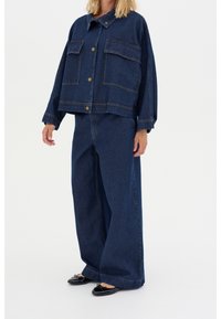 Kvinna som bär en lös, mörkblå denimjacka och vida jeans tillsammans med svarta ballerina skor mot en vit bakgrund.